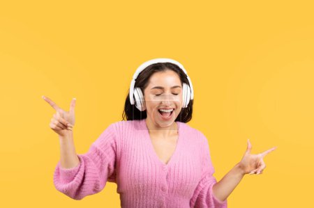 Satisfied young islamic female in wireless earphone has fun, dancing, enjoys free time, listen music, sing, isolated on yellow studio background. Relax, lifestyle, people emotions