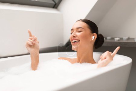 Photo for Relaxing Playlist. Relaxed Lady With Earbuds Listening To Music Online Taking Bath With Foam And Singing Favorite Song In Modern Bathroom Indoors, Posing With Eyes Closed. Domestic Relaxation - Royalty Free Image