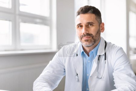 Photo for Professional help. Portrait of confident middle aged physician sitting at desk at his office in clinic and looking at camera. Male doctor wearing stethoscope and white coat - Royalty Free Image