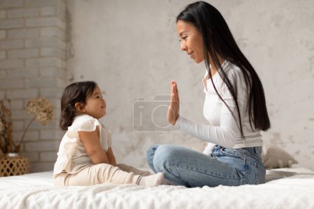 Foto de Maternidad y cuidado de niños. Alegre coreana mamá jugando y aplaudiendo las manos con adorable bebé hija sentado en el dormitorio en casa. Mamá divirtiéndose con su hijo pequeño en interiores. Vista lateral - Imagen libre de derechos