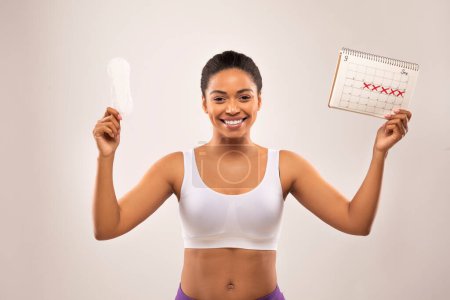 Photo for Happy cheerful young black woman demonstrates periods calendar and sanitary napkin, isolated on grey studio background. Women healthcare, menstruation, menstrual cycle concept - Royalty Free Image