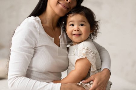 Téléchargez les photos : Coup de feu recadré de maman asiatique embrassant sa fille bébé assis sur le lit à la maison. mignonne petite fille regardant caméra posant avec maman dans chambre moderne. Maternité et baby-sitting Concept - en image libre de droit