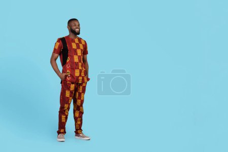 Photo for Handsome black man in traditional african clothes posing over blue studio background, smiling confident young man looking aside at copy space, keeping hands in pockets, full length shot - Royalty Free Image