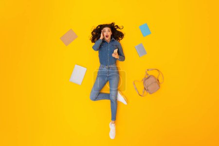 Photo for Shocked Woman Using Smartphone With Educational Application Looking At Camera Lying Among Copybooks And Backpack Over Yellow Studio Background. E-Learning App Concept. Top View Shot - Royalty Free Image