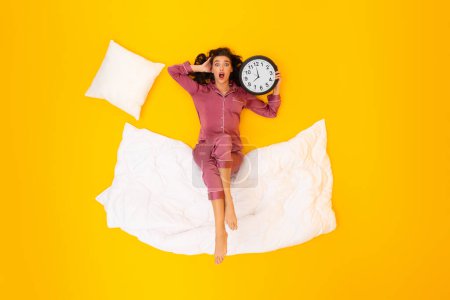 Photo for Oversleeping. Shocked Lady Holding Round Alarm Clock Looking At Camera In Shock Waking Up Lying On Blanket In Studio On Yellow Background. Time Management Problems. Above View Shot - Royalty Free Image