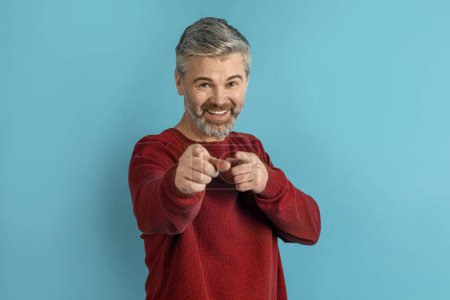 Photo for Hey you. Happy playful handsome grey-haired middle aged man in casual pointing at camera and smiling, isolated on blue studio background, flirting, enjoying good day, copy space - Royalty Free Image