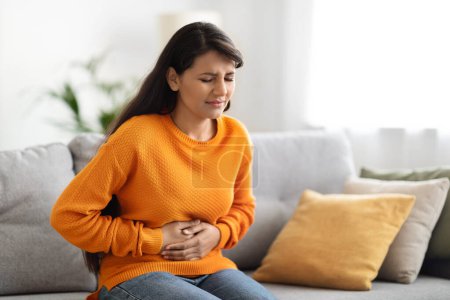 Photo for Concerned unhappy young indian woman in casual suffering from stomach-ache, sitting on sofa at home. Female holding tummy, undergoing belly pain and discomfort, suffer from menstruation, copy space - Royalty Free Image