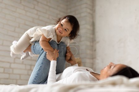 Photo for Happy Japanese Mom Riding Baby Daughter On Legs Lifting Her In Air Having Fun And Playing Lying On Bed At Home. Mother Bonding And Cuddling With Her Adorable Toddler Child. Selective Focus - Royalty Free Image