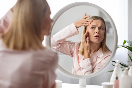 Upset middle aged woman checking wrinkles around her eyes while making beauty routine at home, beautiful mature lady looking in mirror and touching face, feeling tired and stressed, selective focus