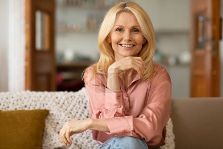 Photo for Happy Blonde Mature Lady Smiling To Camera Holding Hand Near Face Sitting On Sofa At Home. Stylish Attractive Middle Aged Woman Posing In Modern Living Room. Beauty In Older Age Concept - Royalty Free Image