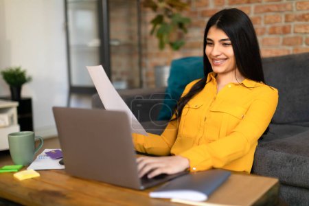 Photo for Happy spanish female student sitting at table with laptop and notepads, smiling and holding sheet of paper, doing homework or writing coursework, copy space - Royalty Free Image