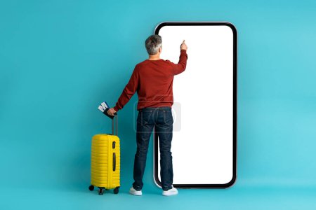 Photo for Checkin online. Middle aged man in casual outfit traveller using huge smatphone with white empty screen, carrying yellow luggage, holding passport with tickets, mockup, blue studio background - Royalty Free Image