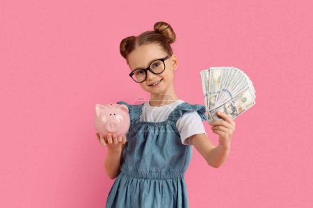 Photo for Investments Concept. Smiling Little Girl Holding Piggybank And Dollar Cash In Hands, Cute Preteen Female Kid Wearing Eyeglasses Enjoying Saving Money, Standing On Pink Studio Background, Free Space - Royalty Free Image