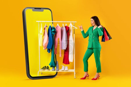 Photo for Stylish lady standing near huge cellphone with blank screen and choosing clothes on rail over yellow studio background, full length. Great mobile offer and application ad - Royalty Free Image