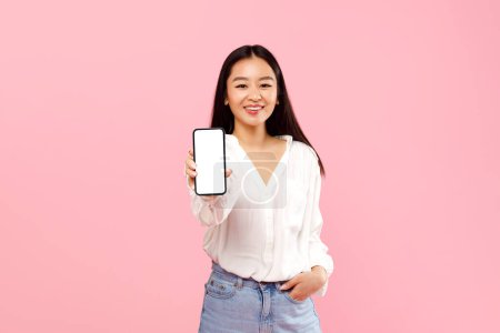 Foto de Anuncio de aplicación móvil. Feliz señora asiática mostrando blanco pantalla vacía del teléfono inteligente, recomendando aplicación o sitio web sobre fondo de estudio rosa, maqueta. Mira esto, pantalla del teléfono celular - Imagen libre de derechos