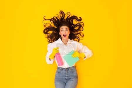 Photo for Shocked Woman Holding Detergent Spray And Rag Advertising Cleaning Products And Cleanup Equipment, Looking At Camera Opening Mouth In Amazement Posing Lying On Yellow Studio Background - Royalty Free Image