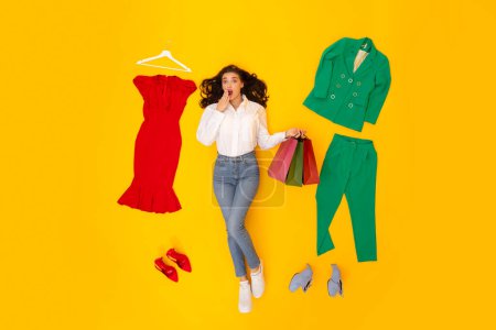 Photo for Fashion And Shopping. Female Stylist Holding Shopper Bags Lying Among Clothes And Shoes Looking At Camera And Covering Mouth In Shock Over Yellow Studio Background. Above View Shot - Royalty Free Image