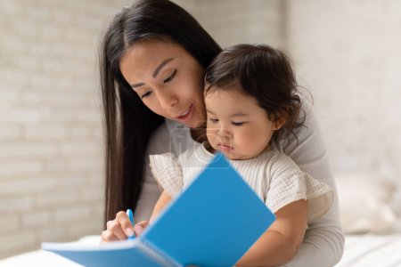 Foto de Mamá china y su hija bebé aprendiendo y dibujando en un cuaderno sentado en la cama en casa. Niñera enseñando a un niño lindo a escribir y dibujar. Concepto de niño inteligente - Imagen libre de derechos