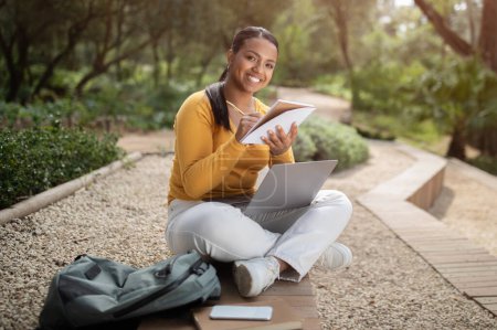 Téléchargez les photos : E-learning. Joyeuse étudiante brésilienne préparant la leçon, utilisant un ordinateur portable et prenant des notes, assise dehors dans le parc. Concept moderne d'enseignement à distance - en image libre de droit