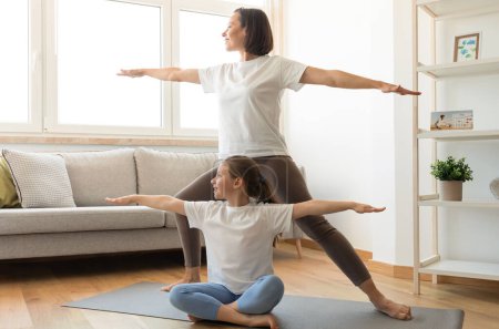 Téléchargez les photos : Mode de vie actif, motivant. Mère millénaire européenne souriante et adolescente dans le yoga de pratique de vêtements de sport, faire de l'exercice, profiter de l'entraînement dans le salon intérieur. Soins du corps et fitness à la maison - en image libre de droit