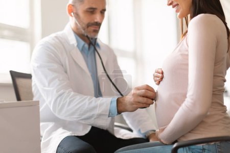 Téléchargez les photos : Gynécologue masculin d'âge moyen avec stéthoscope examinant le ventre de la patiente enceinte, écoutant les battements de cœur des bébés tout en faisant le bilan de santé de la femme enceinte à la clinique - en image libre de droit