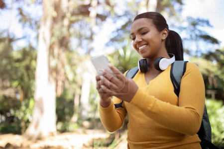 Photo for Happy female brazilian student with smartphone and backpack walking in park, using phone and chatting in social networks, looking at screen and smiling, free space - Royalty Free Image
