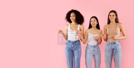 Photo for Look there. Three happy diverse women pointing fingers aside at free space, advertising something, standing over pink background, studio shot, panorama - Royalty Free Image