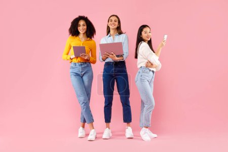 Photo for Three diverse ladies using different gadgets, networking online and browsing internet, standing on pink background, full length. Happy women posing with smartphone, laptop and digital tablet - Royalty Free Image