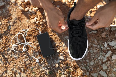 Photo for Millennial african american man tying shoelaces on sneakers on road or beach with headphones, phone with empty screen, unrecognizable. Workout morning, app for body care outdoor, ad, offer - Royalty Free Image
