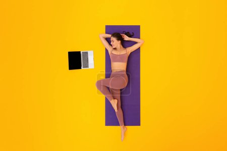 Photo for Online Fitness Workout. Top View Of Fit Lady Exercising Near Laptop Computer Lying On Mat Over Yellow Studio Background, Wearing Fitwear. Woman Having Distance Training. Full Length - Royalty Free Image