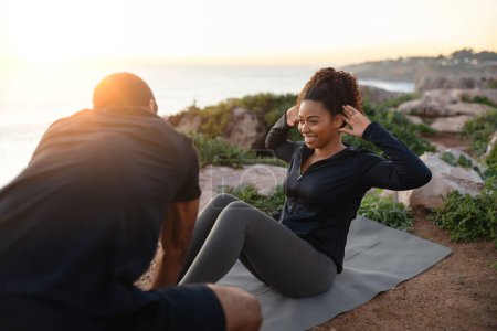 Photo for Smiling millennial african american man help to lady in sportswear do abdominal exercises on ocean beach at sunrise. Support, body care together, workout in morning, lose weight outdoor - Royalty Free Image