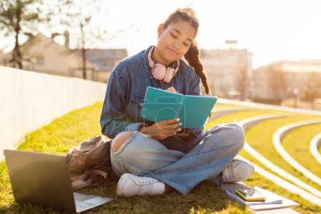 Foto de Lección remota. Estudiante de raza mixta estudiando al aire libre con portátil, sentada en el parque y tomando notas al bloc de notas, disfrutando de la educación en línea, espacio para copiar - Imagen libre de derechos