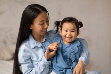 Photo for Portrait Of Cheerful Japanese Mommy And Adorable Baby Daughter Wearing Jeans Clothes Posing Smiling To Camera In Modern Bedroom At Home. Babysitting And Child Care Concept - Royalty Free Image