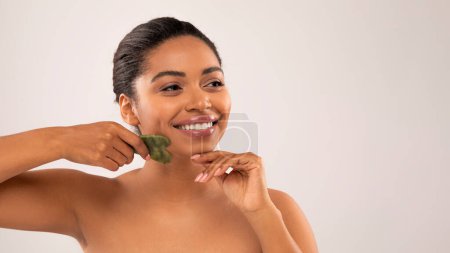 Téléchargez les photos : Joyeux sourire joyeux attrayant moitié nue jeune femme noire faisant massage du visage avec gua sha dans la routine de soins du visage du matin, en regardant l'espace de copie, gros plan, fond de studio, bannière - en image libre de droit