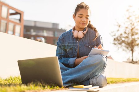 Photo for Exam study concept. Black teen student girl taking notes while watching online lesson on laptop computer, sitting outdoors in park, copy space - Royalty Free Image