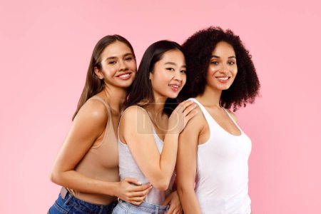 Photo for Three beautiful multiracial millennial women embracing and smiling to camera, standing on pink background, studio shot. Diverse female beauty - Royalty Free Image