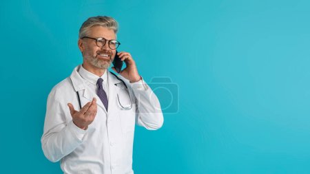 Photo for Happy handsome middle aged grey-haired doctor have phone call with patient, consulting remotely, smiling and gesturing over blue studio background, looking at copy space, panorama - Royalty Free Image