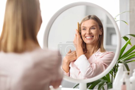 Téléchargez les photos : La routine beauté. attrayant moyen âge femme peigner ses beaux cheveux blonds avec pinceau tout en étant assis près du miroir dans la chambre à coucher, heureuse dame mature regardant à son reflet et souriant - en image libre de droit