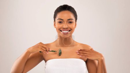 Téléchargez les photos : Joyeux sourire sensuel attrayant jeune femme noire enveloppée dans une serviette dorloter son beau corps après la douche, massage poitrine avec rouleau en pierre de jade, fond de studio, panorama - en image libre de droit