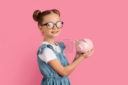 Photo for Wise Saving. Portrait Of Cute Little Girl In Eyeglasses With Piggy Bank In Hands Posing Over Pink Studio Background, Smiling Preteen Female Child Enjoying Economy, Collecting Pocket Money - Royalty Free Image