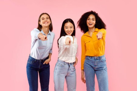 Téléchargez les photos : Hé, toi ! Portrait de femmes multiethniques heureuses pointant du doigt la caméra et souriant, posant sur fond rose, prise de vue en studio - en image libre de droit