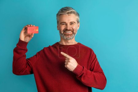 Photo for Cheerful handsome grey-haired mature man in casual pointing at red plastic bank card and smiling, recommending contactless payment, easy fast banking, blue studio background, copy space - Royalty Free Image