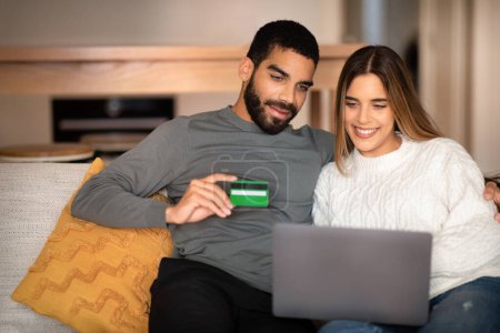 Photo for Positive millennial arabic husband hugs caucasian wife, uses laptop and credit card, enjoy online shopping in living room interior. Sale, order purchase with device, cashback at home - Royalty Free Image