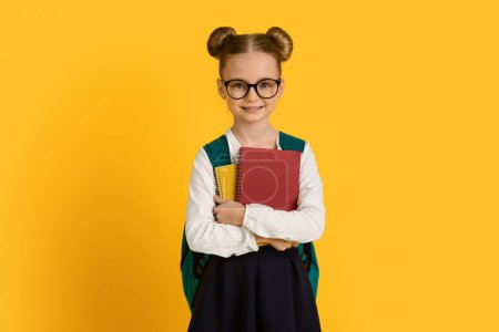 Téléchargez les photos : Belle petite écolière portant des lunettes de vue tenant des livres et souriant à la caméra tout en posant sur fond jaune studio, mignon Preteen enfant féminin bénéficiant d'étude et d'éducation, Copier l'espace - en image libre de droit