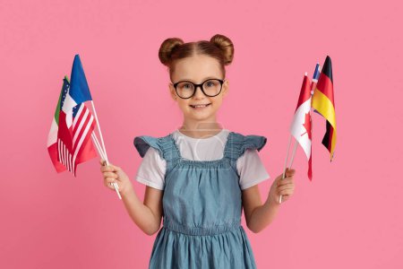 Photo for Cute little girl holding bunch of internetional flags and smiling at camera, happy preteen female child wearing eyeglasses enjoying study foreign languages, standing on pink background, copy space - Royalty Free Image