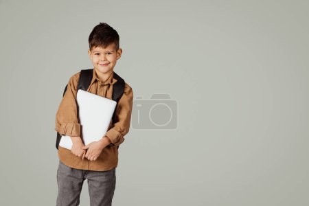 Téléchargez les photos : Joyeux petit enfant européen de 6 ans élève décontracté avec sac à dos et ordinateur portable isolé sur fond gris, studio. Enfance, connaissance, éducation et études à l'école primaire avec dispositif - en image libre de droit