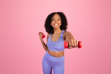 Téléchargez les photos : Femme afro-américaine millénaire souriante en tenue de sport faisant des exercices manuels avec des haltères isolés sur fond de studio rose. Entraînement musculaire, sport, perte de poids et soins du corps, annonce, offre - en image libre de droit