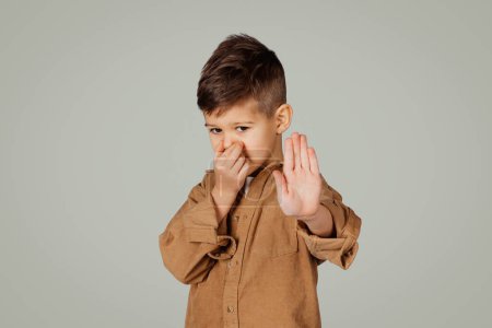 Téléchargez les photos : Désespéré petit enfant de 6 ans dans occasionnel fait geste d'arrêt avec les mains, ferme son nez isolé sur fond gris. Odeur, mauvaise odeur, enfance, étude, émotions dégoûtantes - en image libre de droit