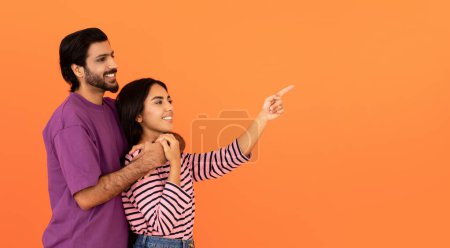 Téléchargez les photos : Amour joyeux beau jeune couple indien dans l'étreinte décontractée, hindou homme et femme regardant et pointant vers l'espace libre pour la publicité sur fond de studio orange, panorama - en image libre de droit