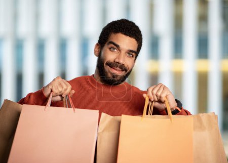 Foto de Compras exitosas. Retrato de un feliz comprador de Oriente Medio sosteniendo bolsas de papel posando al aire libre cerca del centro comercial, sonriendo a la cámara. Concepto de anuncio de venta estacional - Imagen libre de derechos